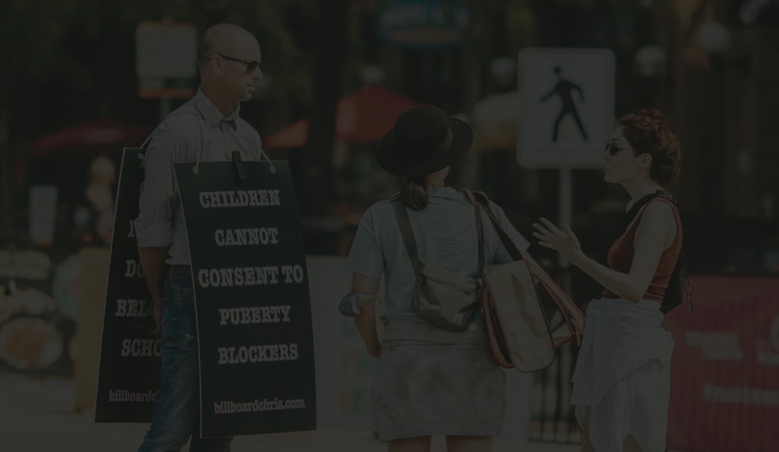 Billboard Chris speaking on a public sidewalk while wearing a sandwich board that reads Children cannot consent to puberty blockers